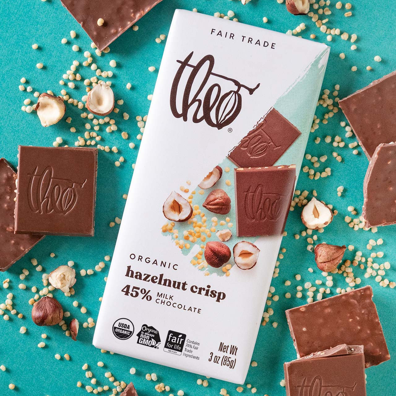Theo Organic Hazelnut Crisp 45% milk chocolate bar packaging with chocolate pieces and nuts on a teal background