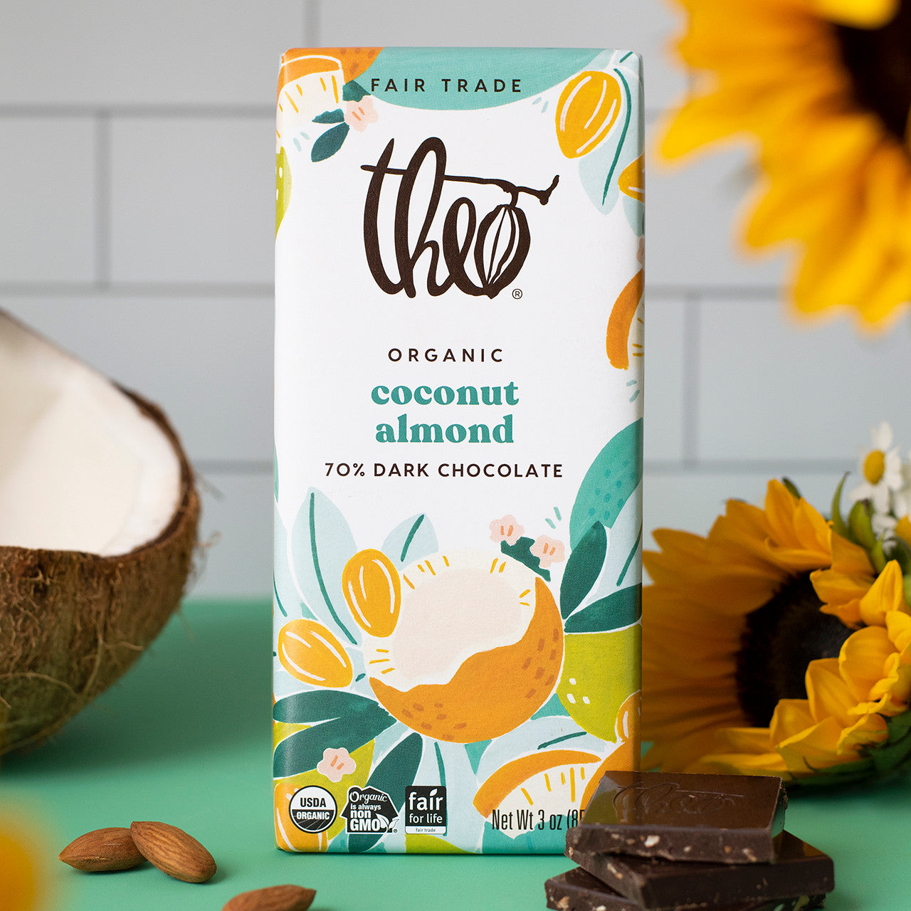Theo Organic Coconut Almond 70% Dark Chocolate bar packaging with a coconut, sunflower, and chocolate pieces on a green surface.