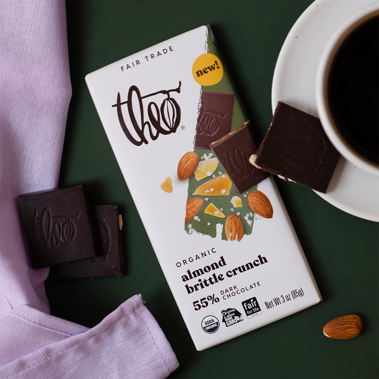 Theo almond brittle crunch dark chocolate bar packaging with a cup of coffee and chocolate pieces on a green surface.