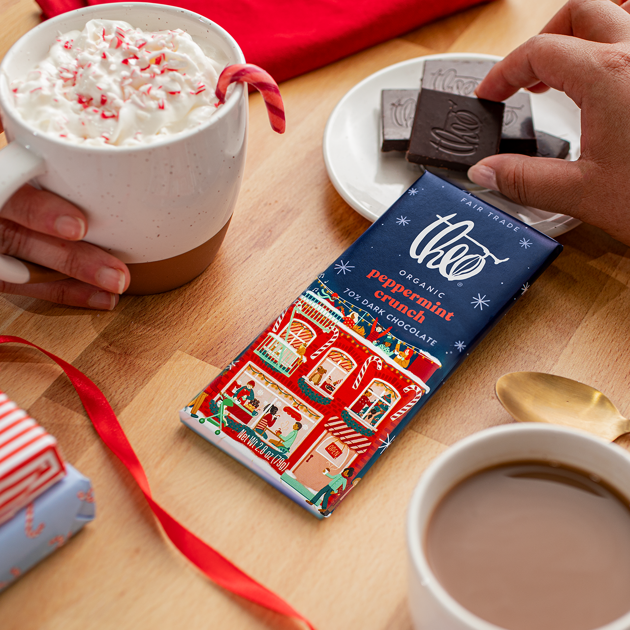 Two cups of hot chocolate with whipped cream and candy canes, a bar of Theo Organic Chocolate on a wooden table.