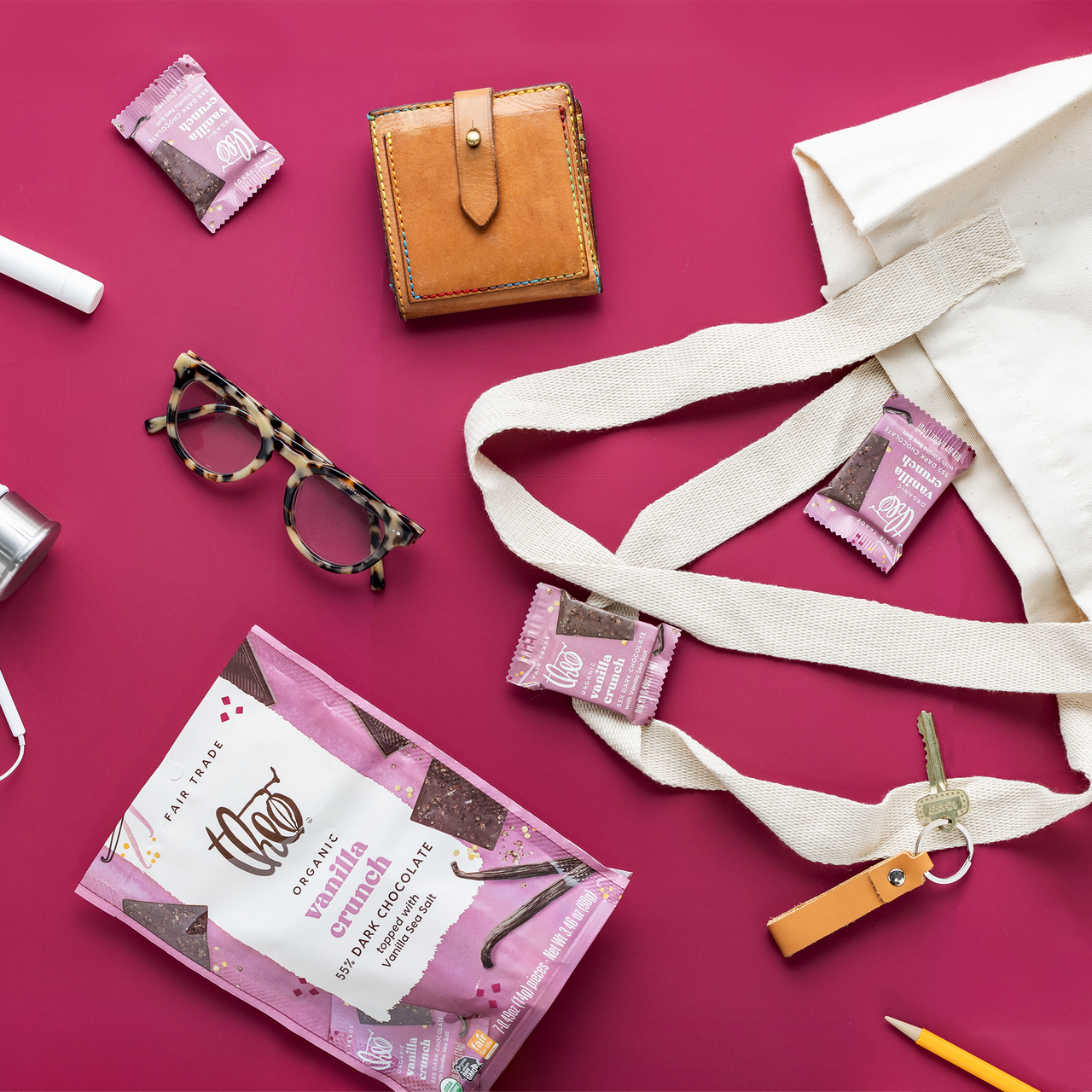 Assorted items including a brown wallet, pink Theo Singles, Organic Theo Vanilla Crunch 55% Dark Chocolate package, and a beige strap on a pink background