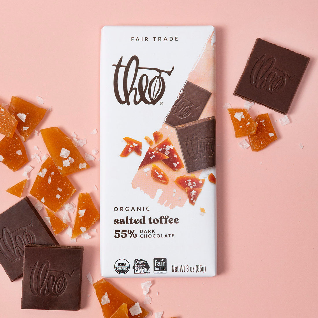 Theo Organic salted toffee 55% dark chocolate bar with chocolate pieces, toffee pieces, and salt on a pink background
