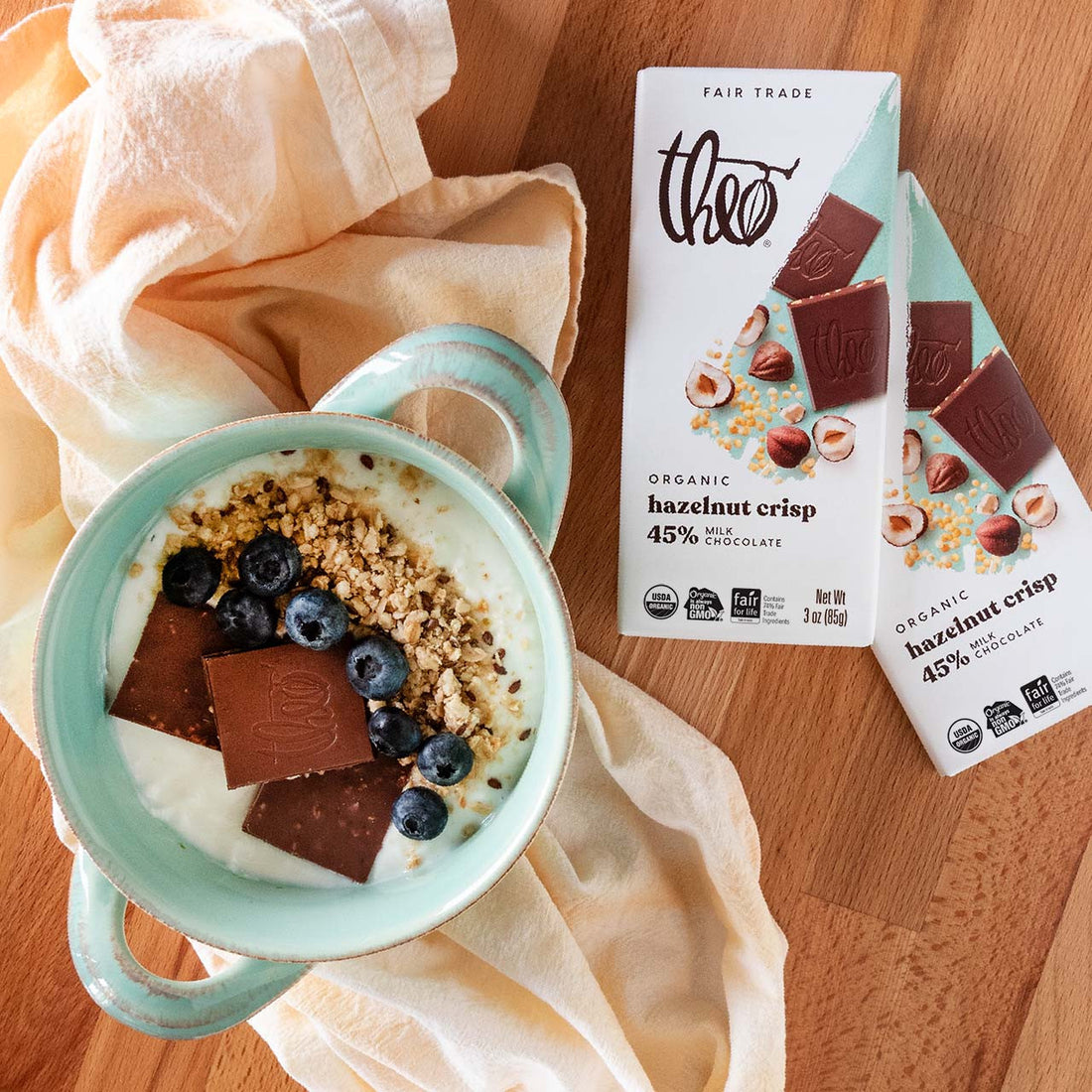 Mint green bowl with yogurt, granola, chocolate, and berries next to a bar of Theo Organic Hazelnut Crisp chocolate on a wooden surface.