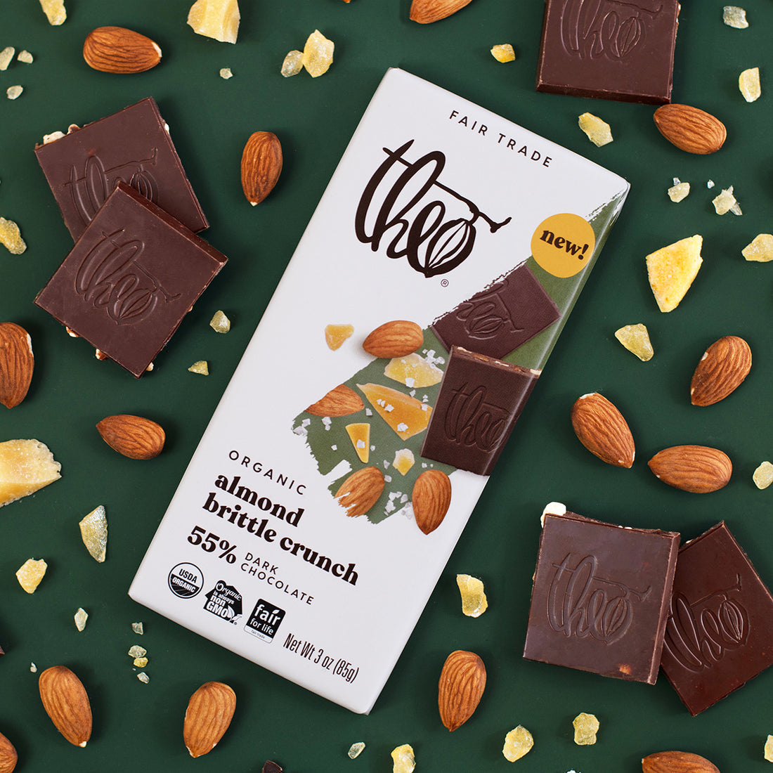Theo Organic Almond Brittle Crunch 55% Dark Chocolate bar on a green background with chocolate pieces and almonds.