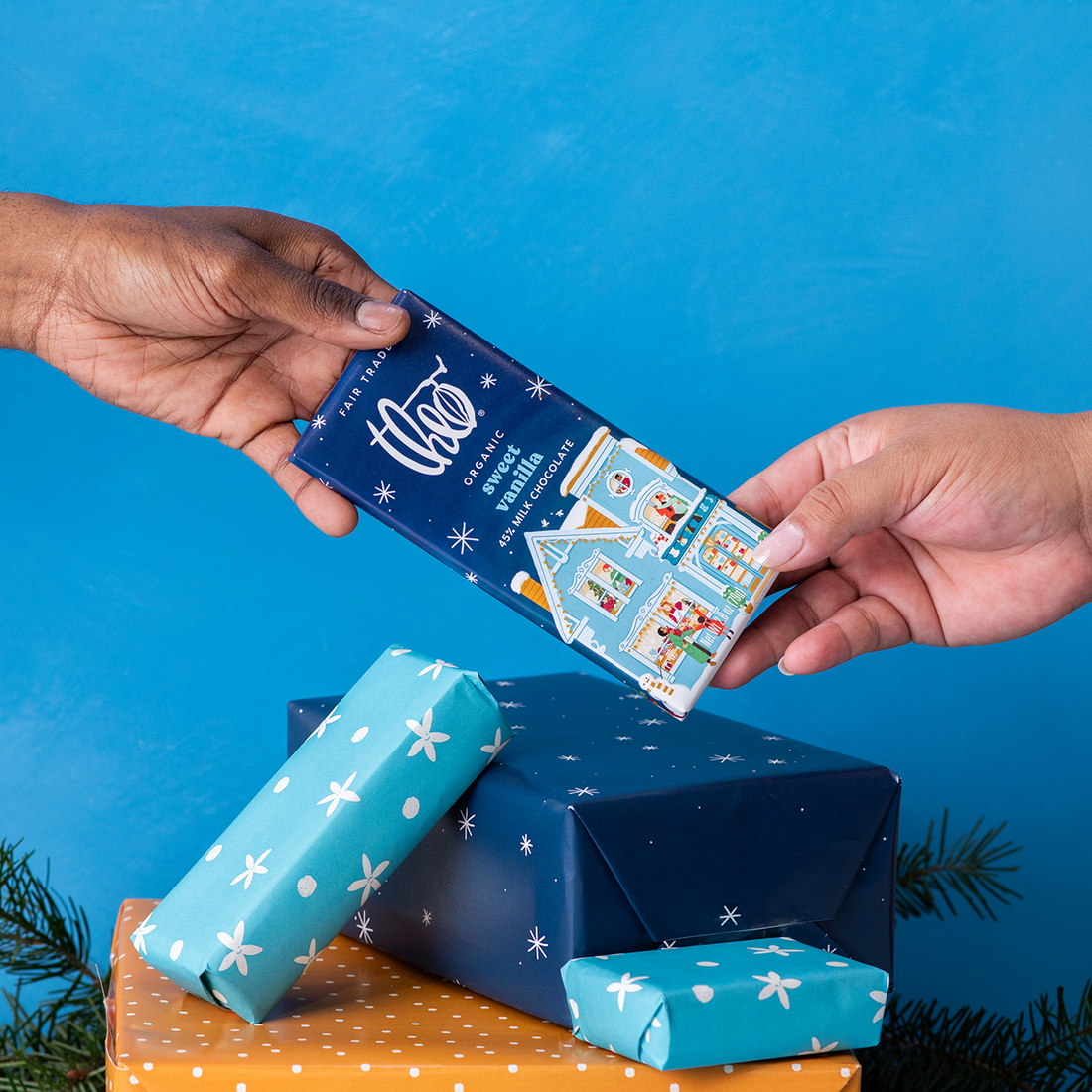 Two hands exchanging a bar of Theo sweet vanilla chocolate against a blue background with wrapped presents.