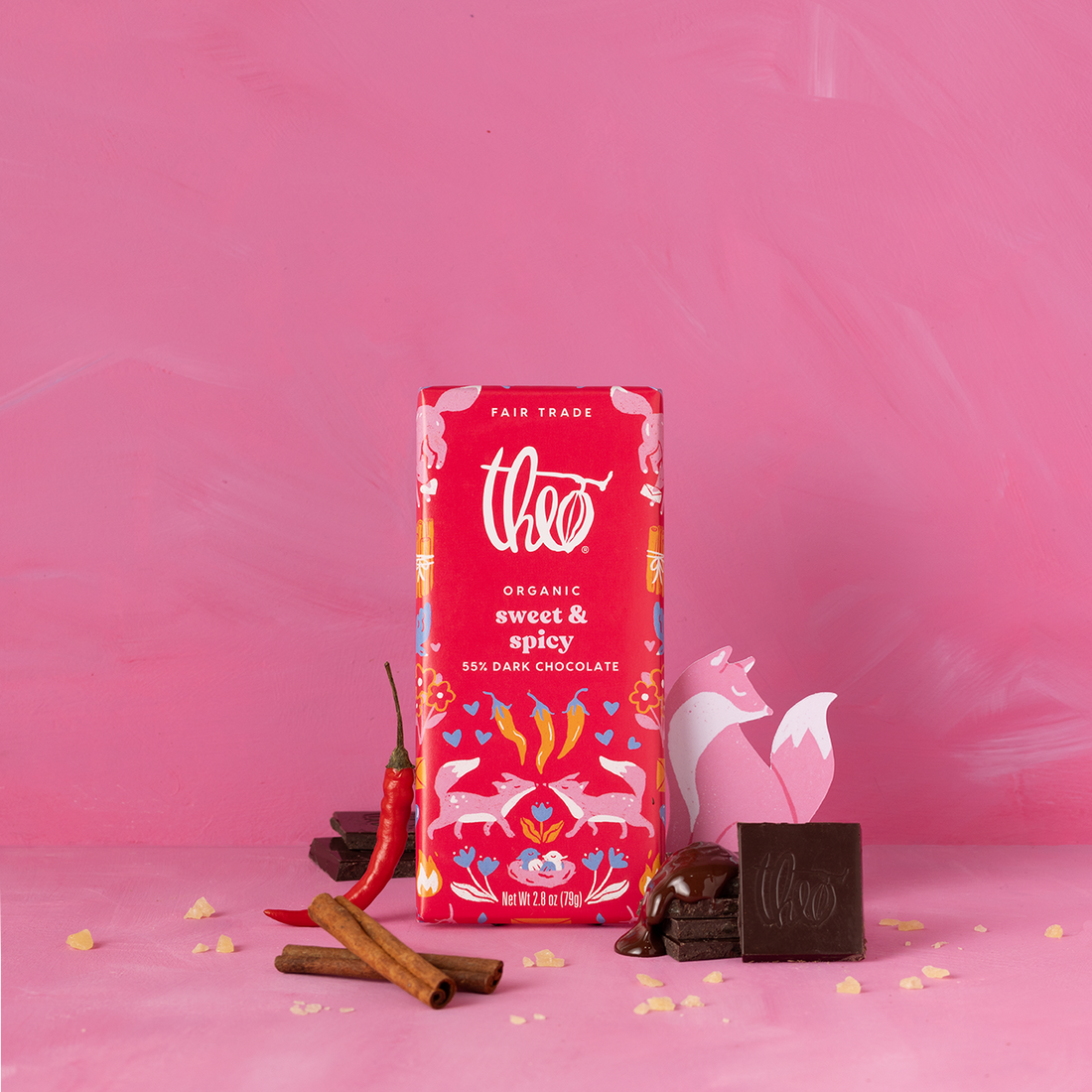 Red package of Theo Organic sweet &amp; spicy dark chocolate on a pink background with cinnamon sticks and chili peppers.