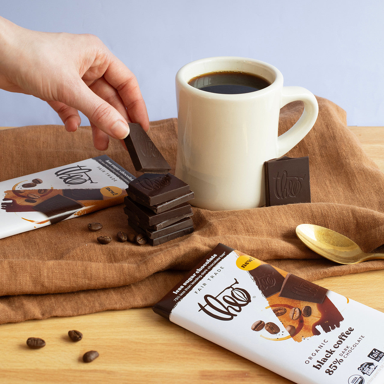 Hand picking a chocolate bar from a stack next to a cup of coffee and Theo Black Coffee 85% dark chocolate packaging on a wooden surface.