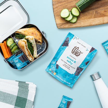Lunchbox with sandwich, vegetables, and a package of Theo Sweet &amp; Salty chocolate singles on a light blue background